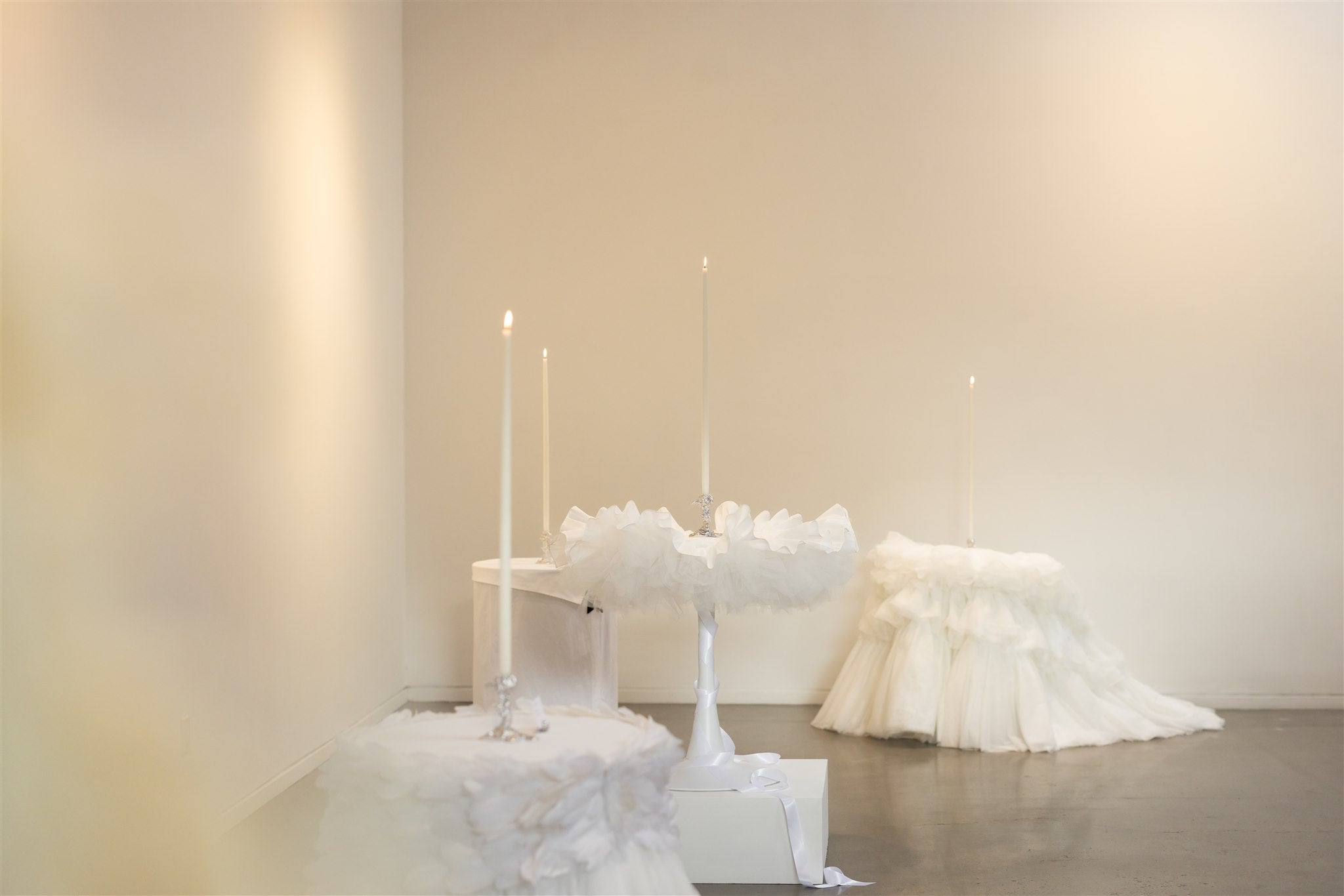 Four bespoke table cloths sit on tables in a large white gallery space, each with a tall lit candle placed in the centre of the tables.