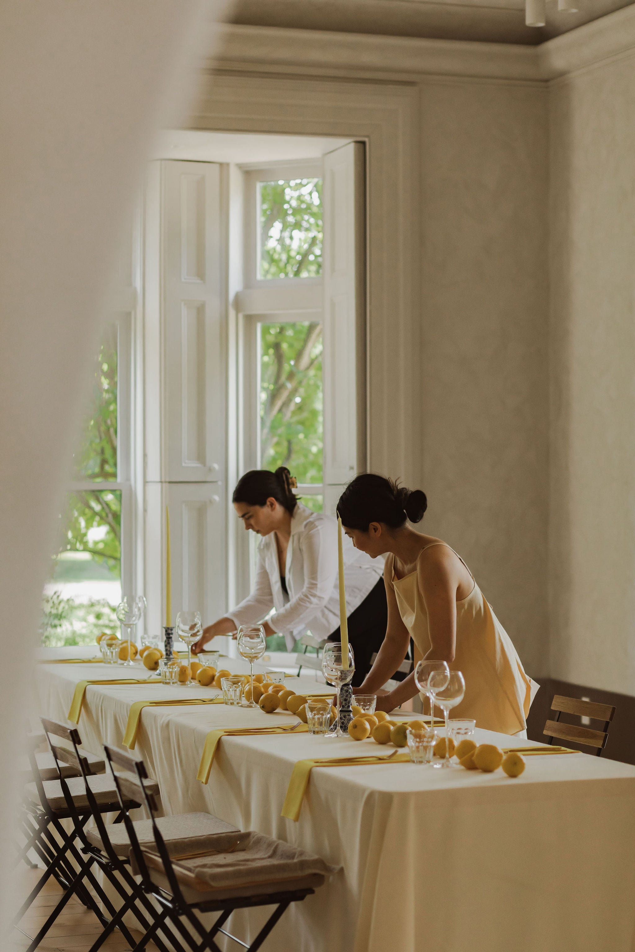 two women set a long table with yellow napkins, tall wineglasses, a long line of lemons runs down the centre of the table