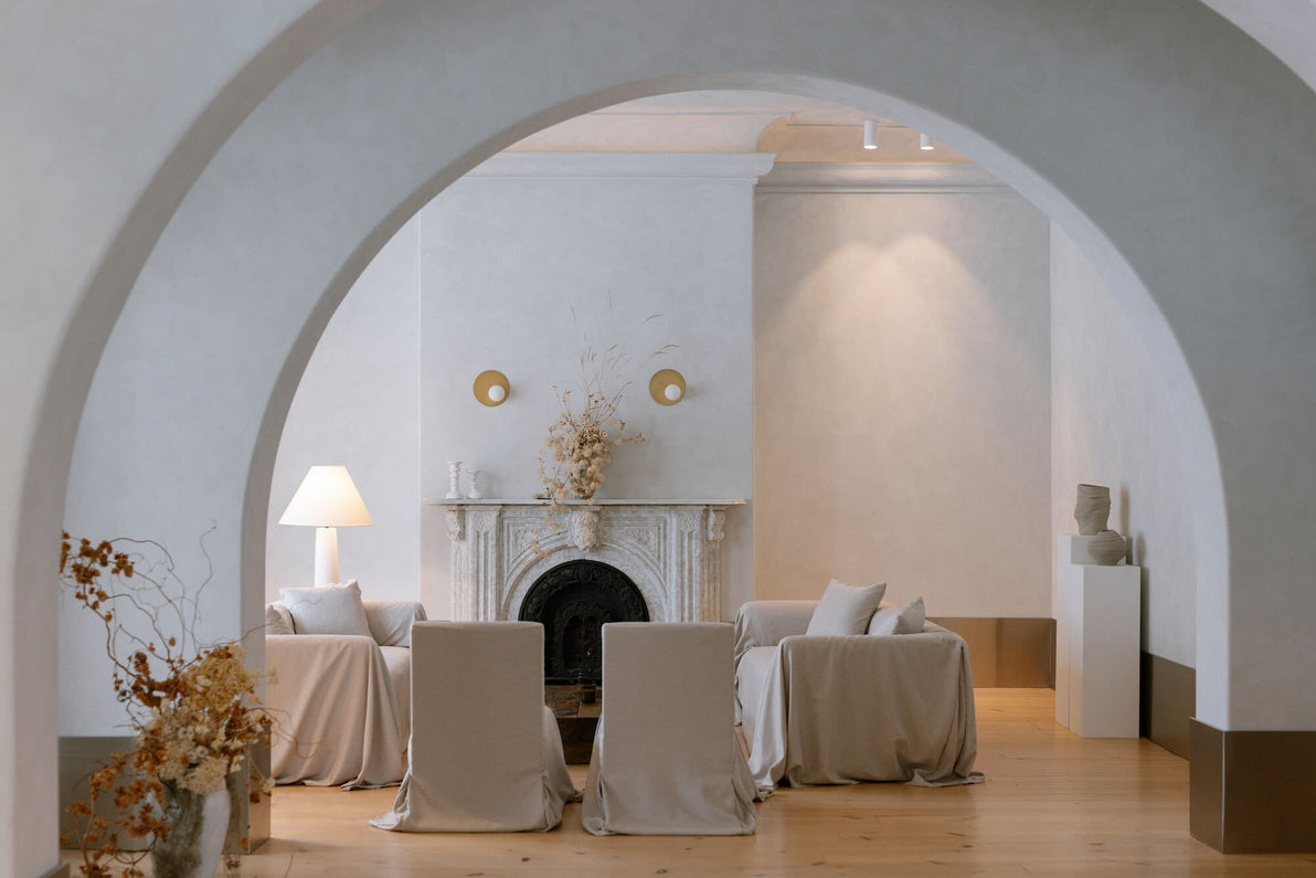 two couches and matching chairs sit in front of a white marble fireplace, warm grey plaster walls, large arches separate the two rooms